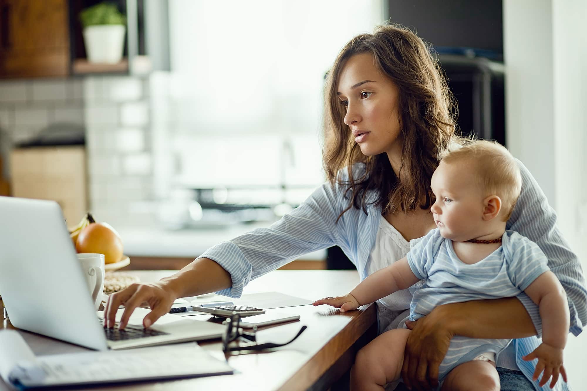 Young woman with baby son working on laptop while planning her home finances.